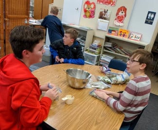 Boys sitting at a table measuring ingredients into a silver bowl